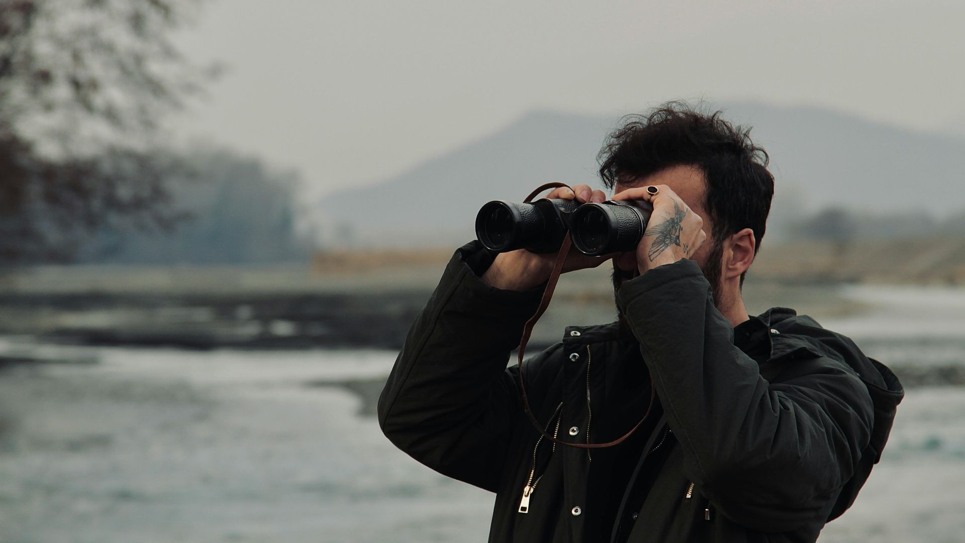A man with tattoos uses binoculars to view a scenic landscape outdoors during the day.