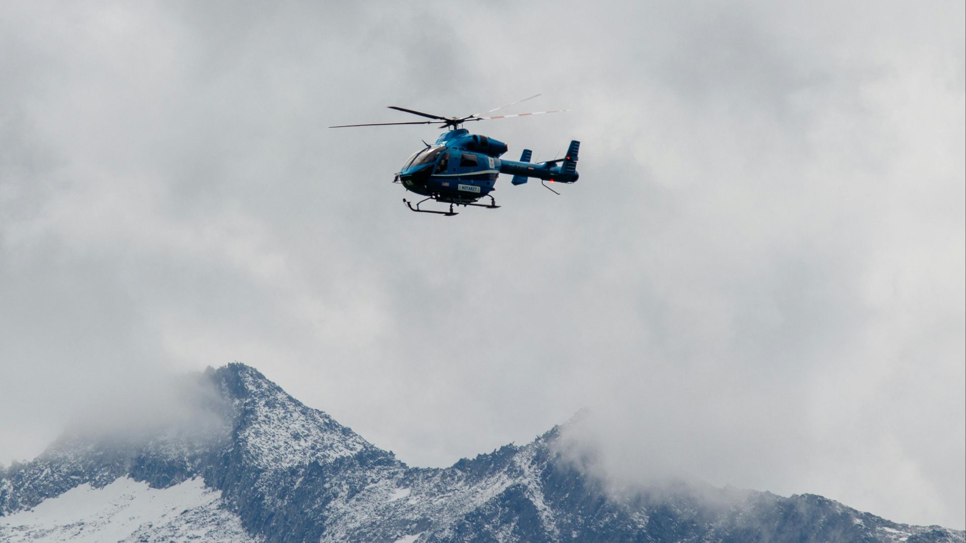A blue helicopter in flight against a backdrop of misty, snow-covered mountains.