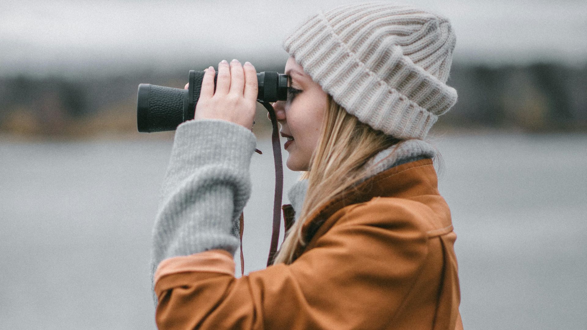 Side view of stylish female in warm woolen hat and coat admiring gray river on blurred background