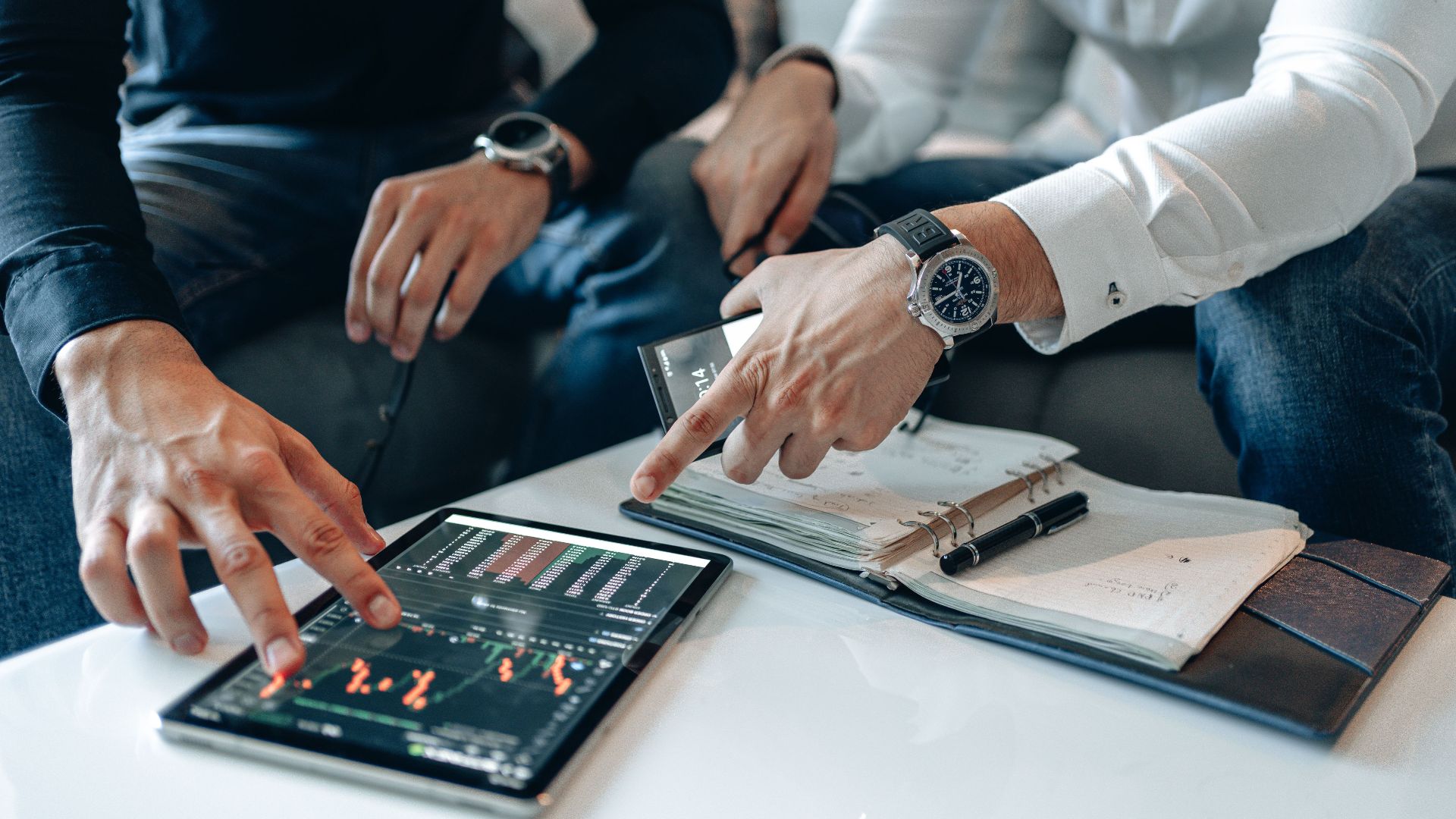 Men in business attire discussing financial data on a tablet indoors.