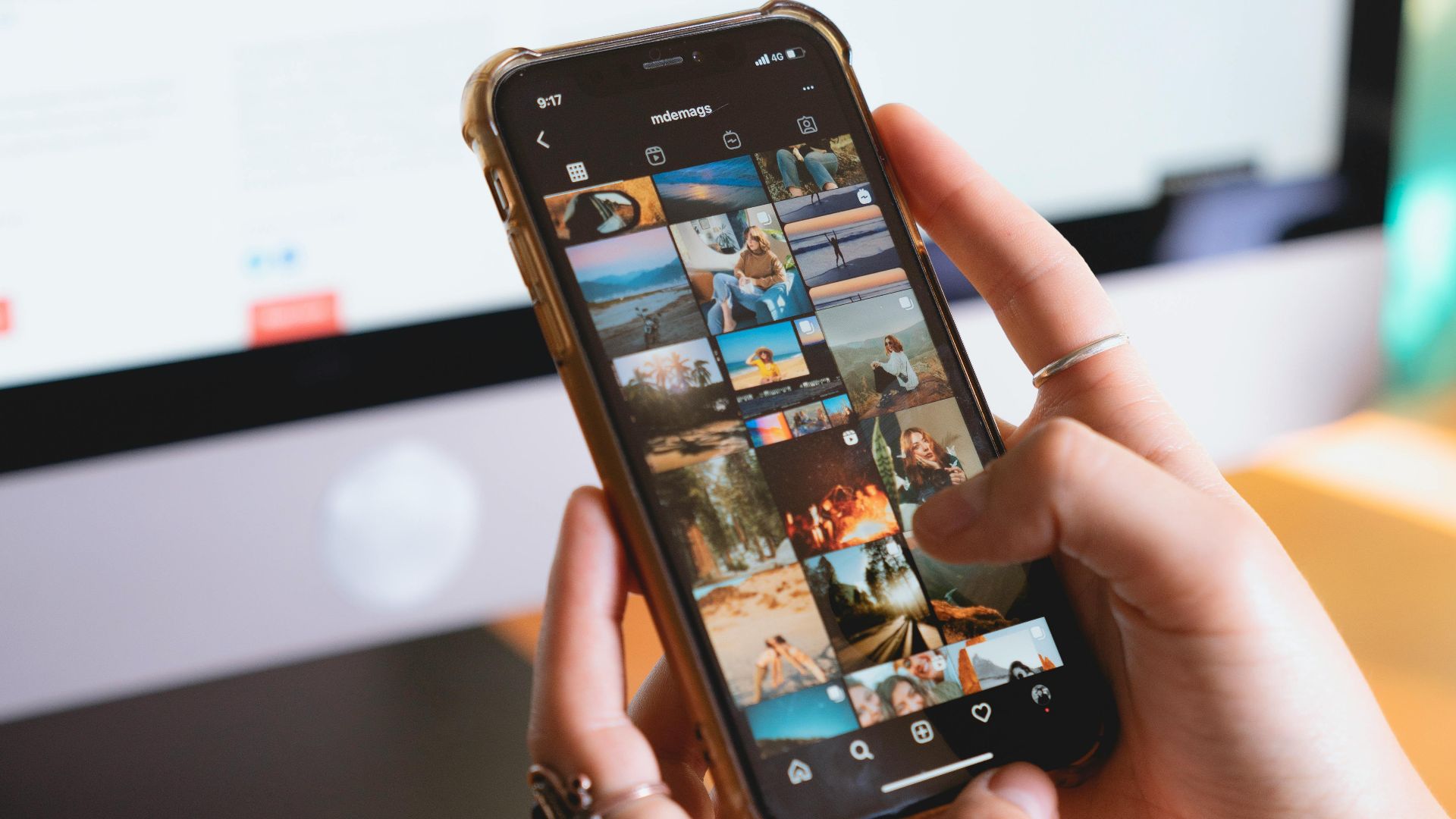 Close-up of a person browsing a smartphone with an Instagram feed, with a computer monitor in the background.