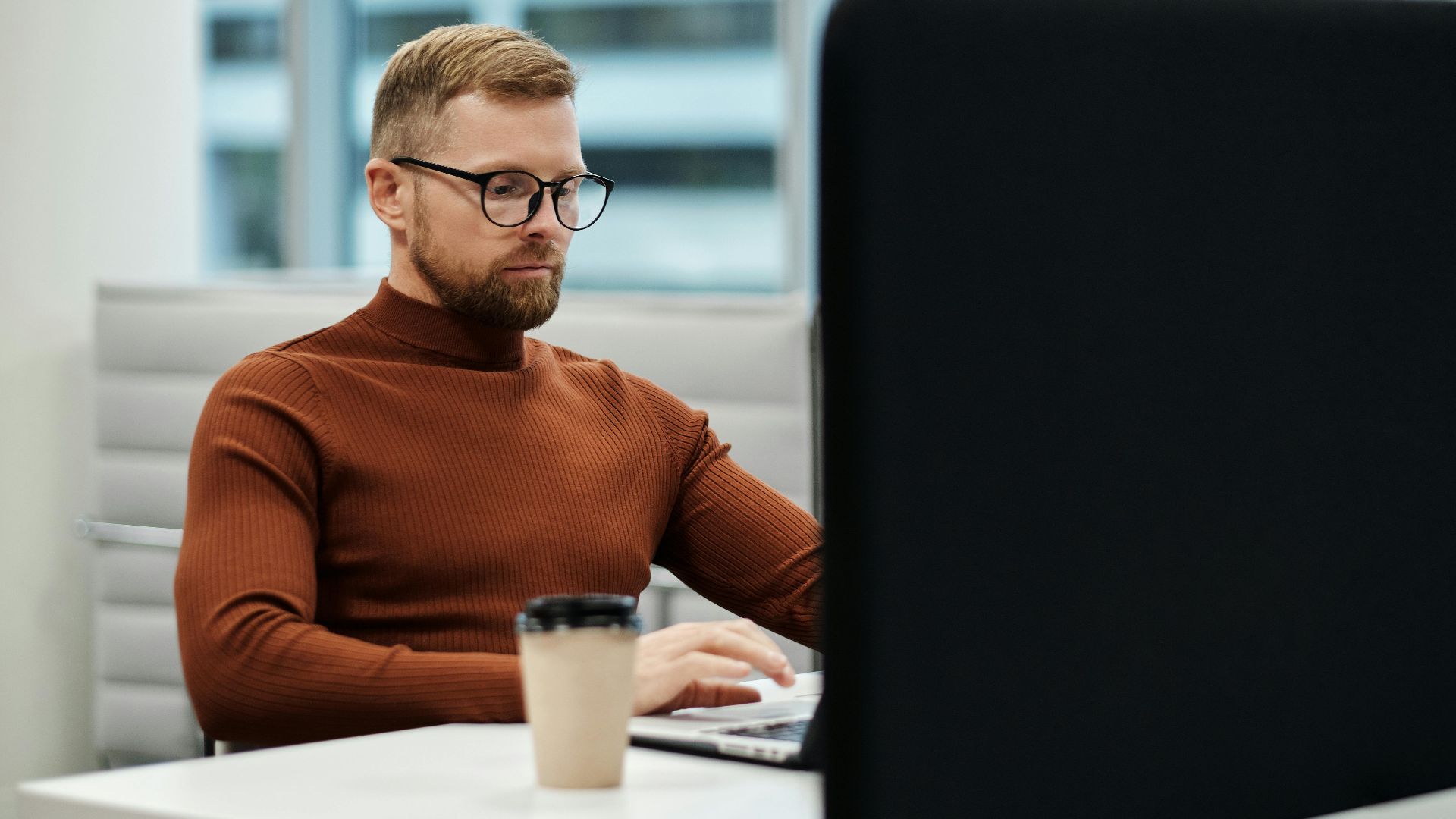 Caucasian man with beard and glasses working at a desk in a modern office environment.