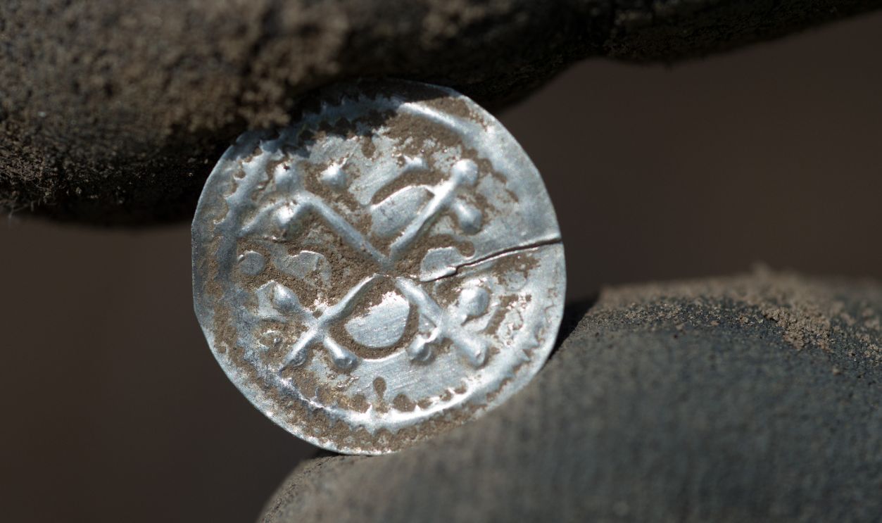 Getty Images - 976862576 - An archeologist holds a cross bracteate minted under Harald Bluetooth (936-984), the first independent Danish coins, after the excavation. Archaeologists have discovered valuable, predominantly Scandinavian silver treasures from the late 10th century, in a field on the German island of Ruegen. In an area of around 400 square metres near the municipality of Schaprode, they recovered jewellery and around 500 - 600 coins, of which more than 100 coins date back to the reign of Danish king Harald "Bluetooth" (910-987).