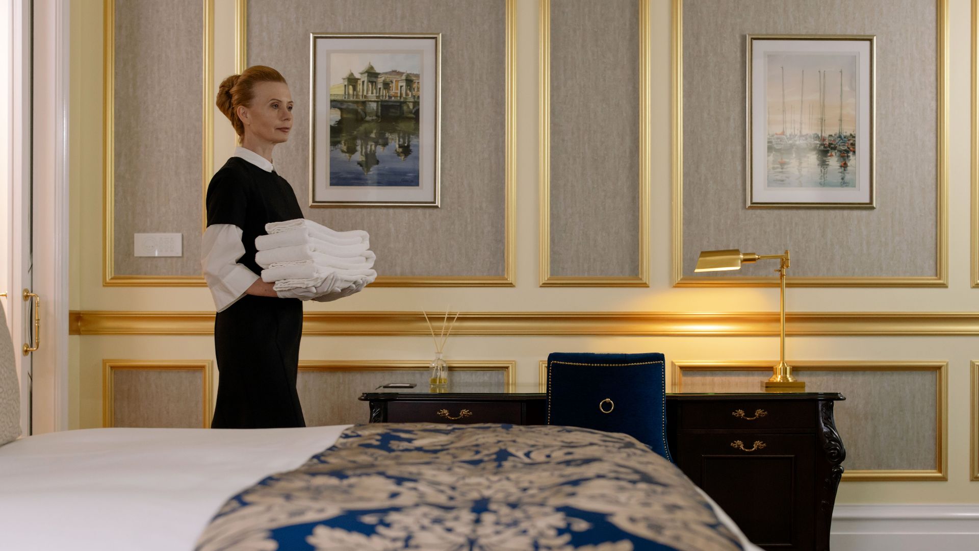 A housekeeper holding towels in a luxurious hotel room with elegant decor and a classic desk.
