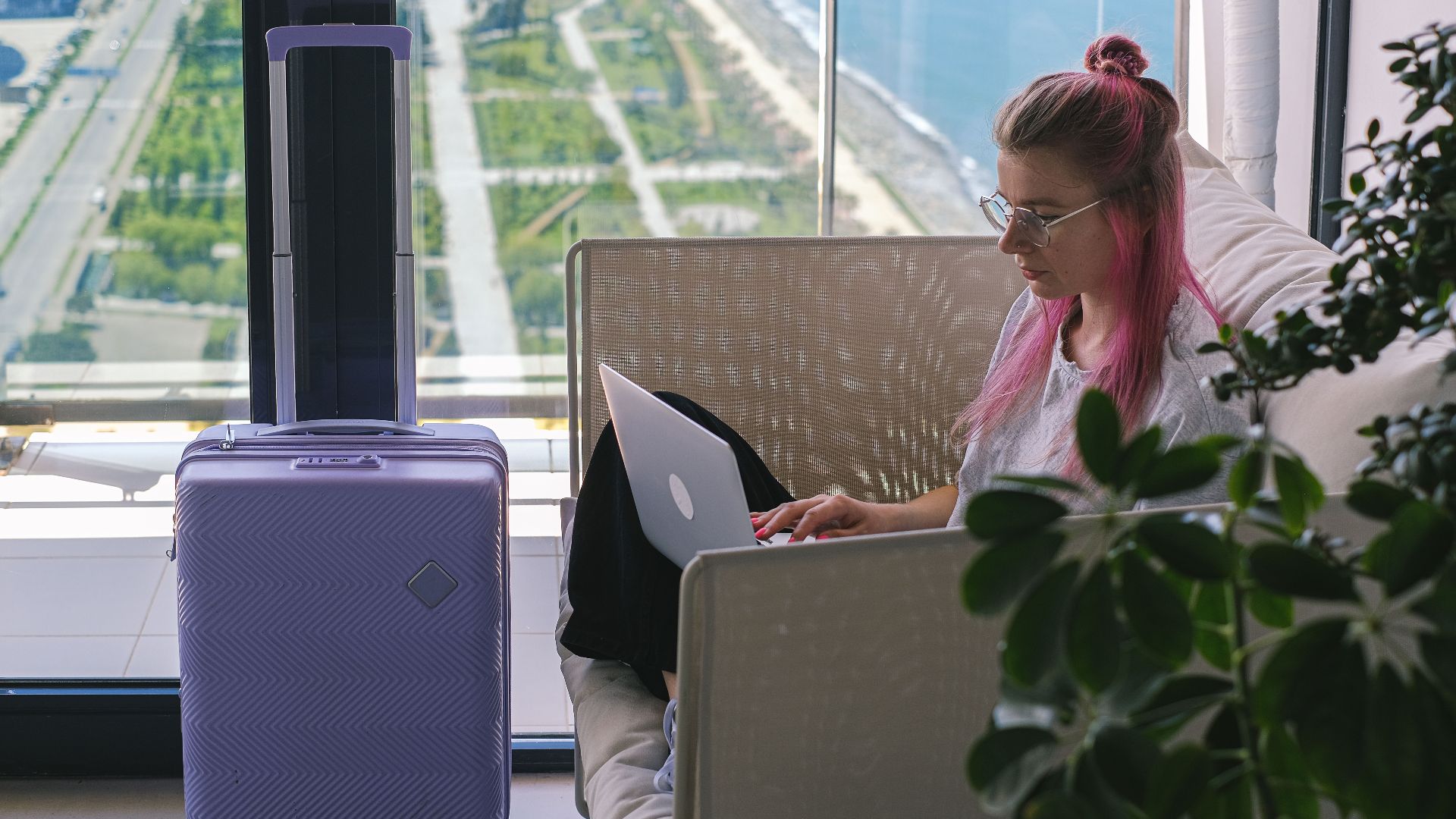 Woman with pink hair working on laptop in hotel room overlooking scenic landscape. Travel and freelance concept.