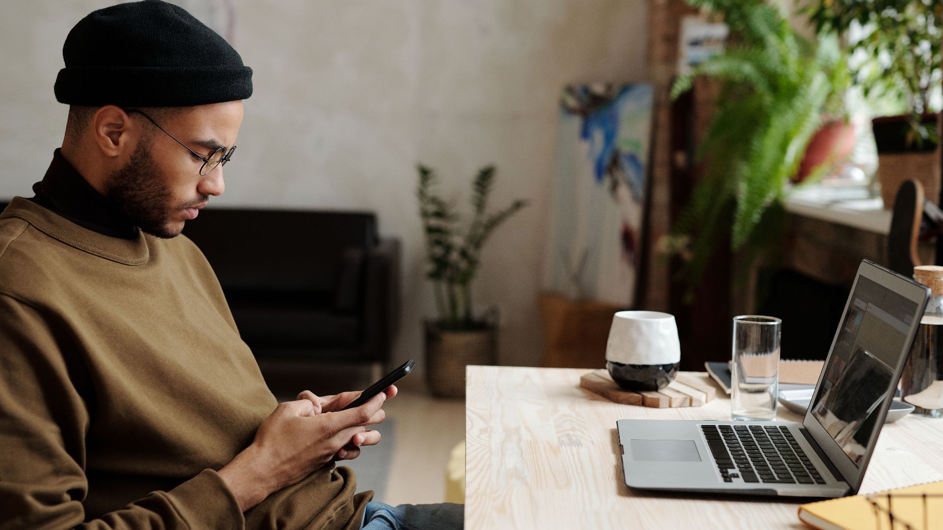 Young man using his phone at a serene home office with a laptop nearby, embodying remote work lifestyle.