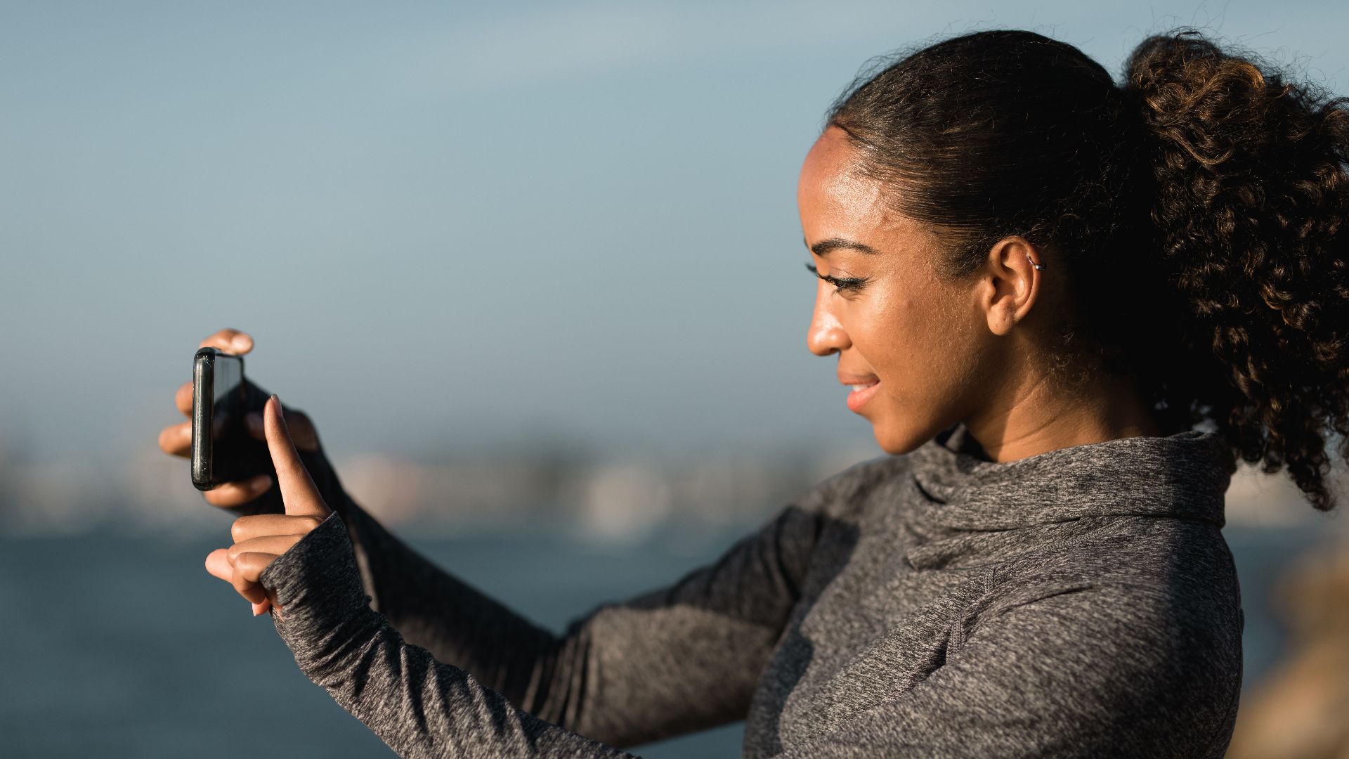 African American woman smiling while taking a photo with her smartphone outdoors near water.