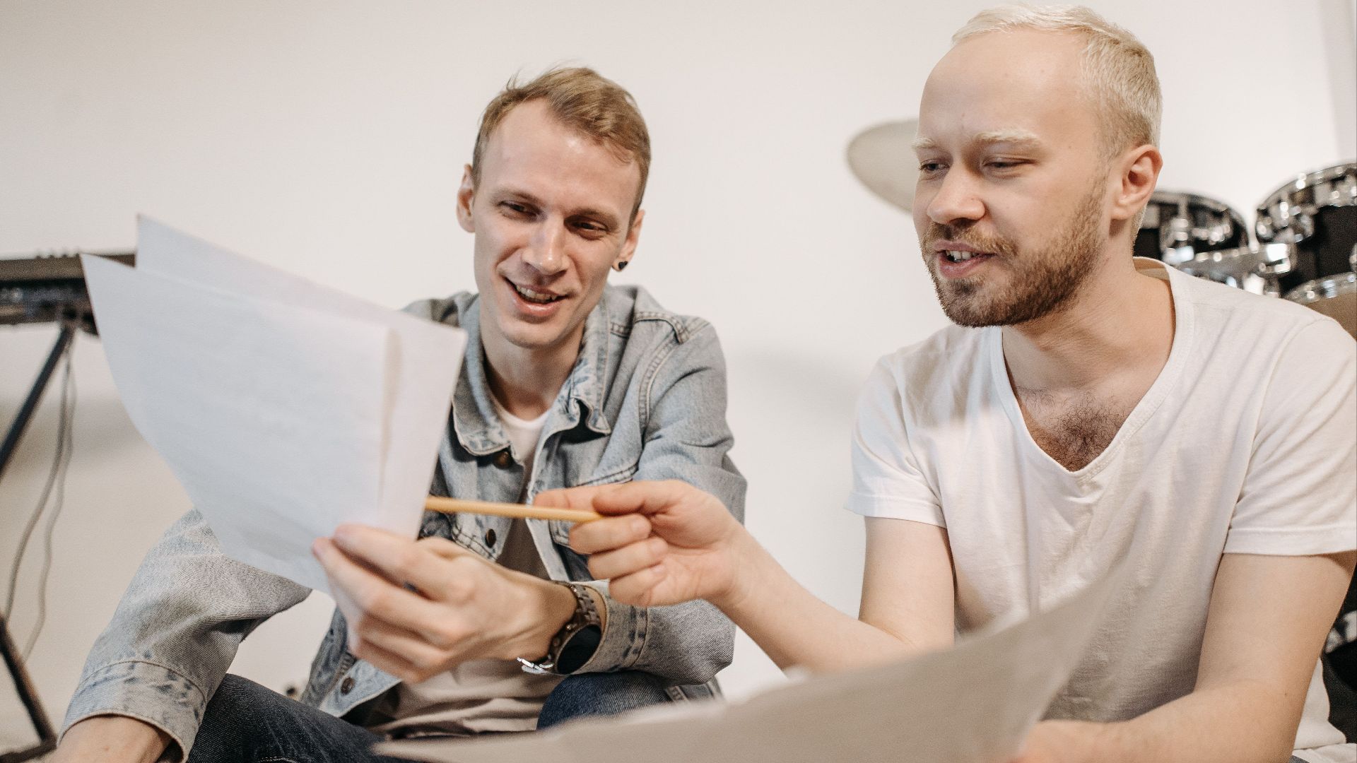 Two men reviewing music sheets with instruments in studio setting.