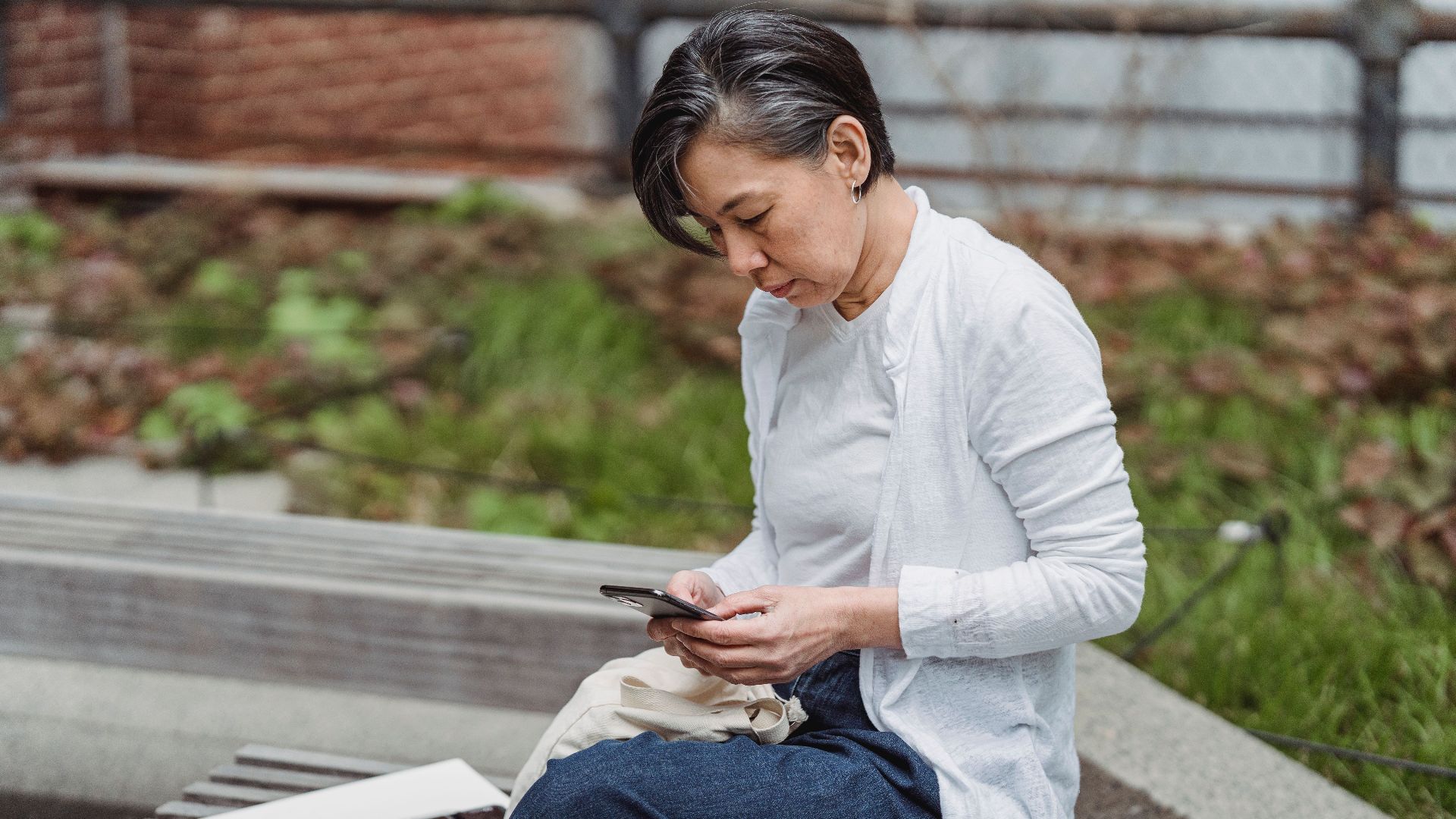 A mature woman in casual attire using her smartphone while sitting on a bench outdoors.