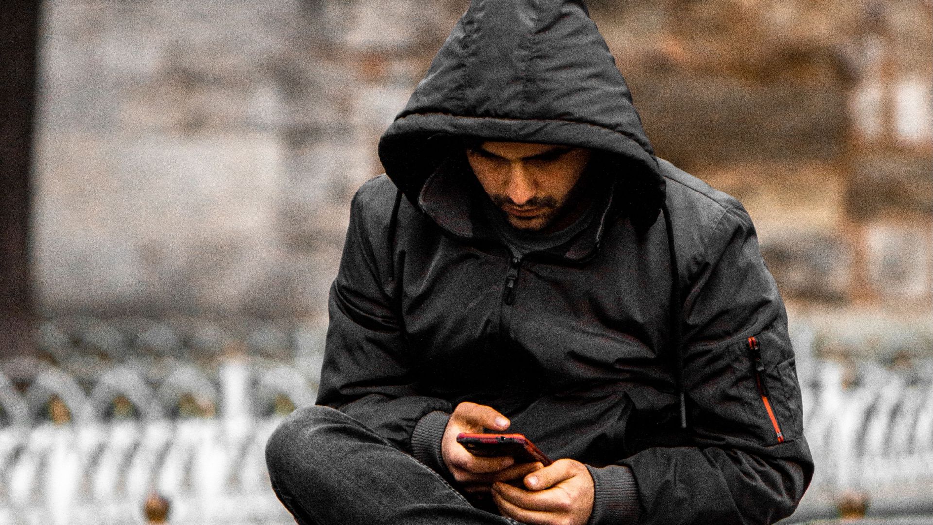 A man wearing a hooded jacket sits on a wooden bench outdoors using his smartphone.