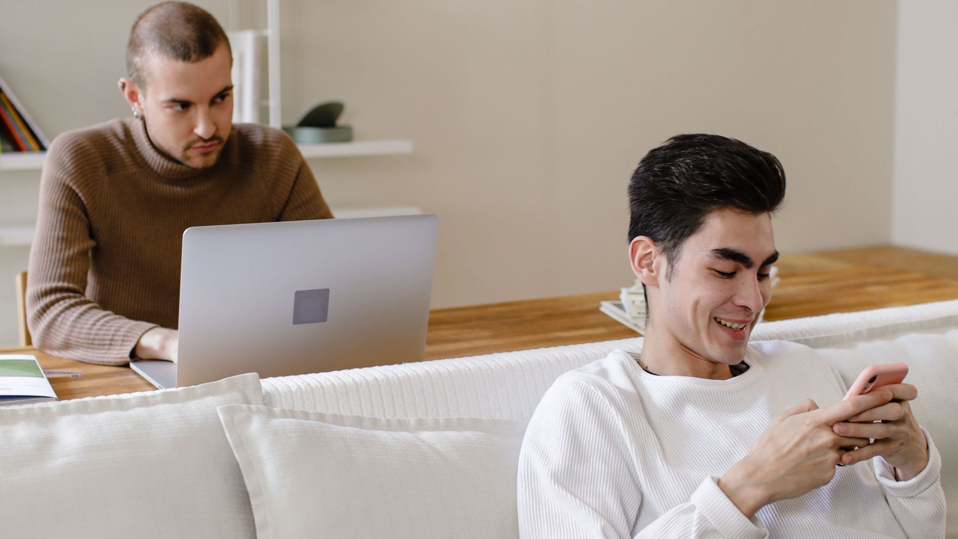 Two men relaxing at home; one working on a laptop, the other on a smartphone, enjoying leisure time.