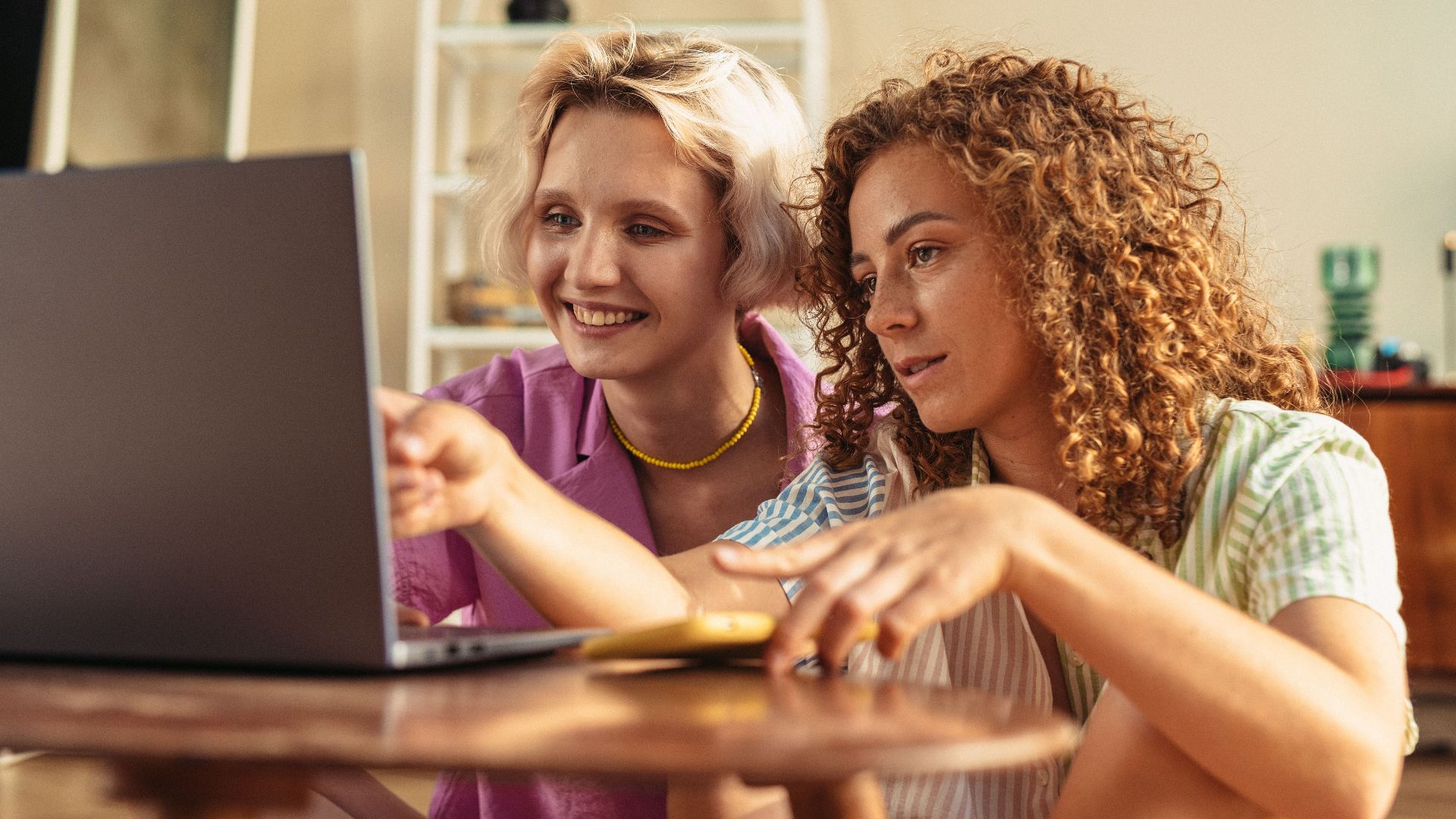 Two women sitting together indoors, sharing a moment as they work on a laptop.