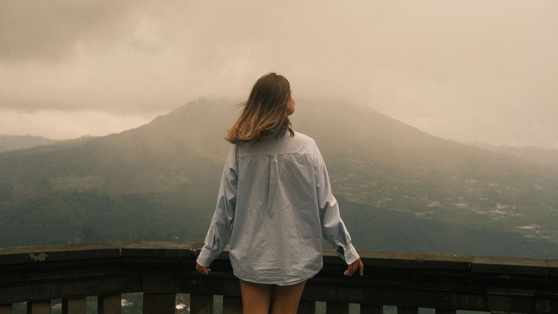 A woman stands on a balcony overlooking a misty mountain landscape during the day.