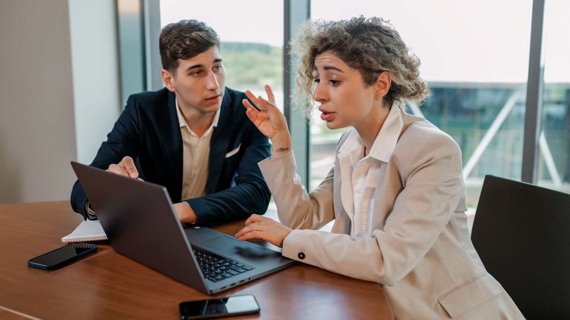 Two business professionals discussing ideas at a wooden table inside a modern office environment.
