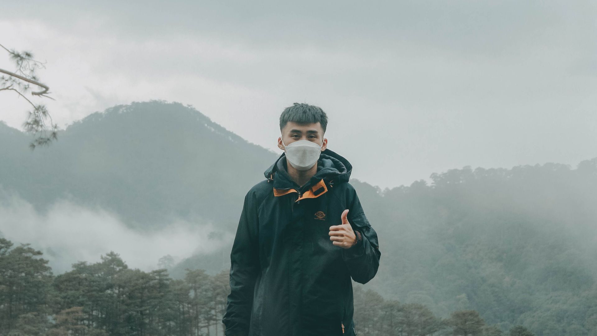 Man wearing a face mask gives a thumbs up while standing outdoors with misty mountains as backdrop.