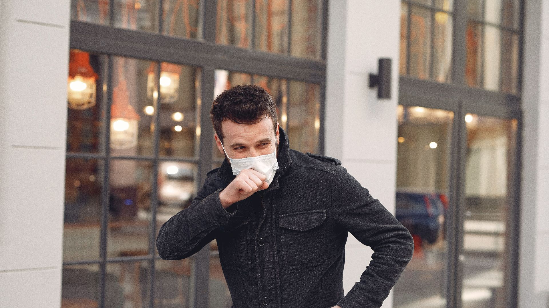 Unhealthy male wearing warm jacket and protective facial mask coughing while standing alone on urban street against city restaurant exterior in cold season