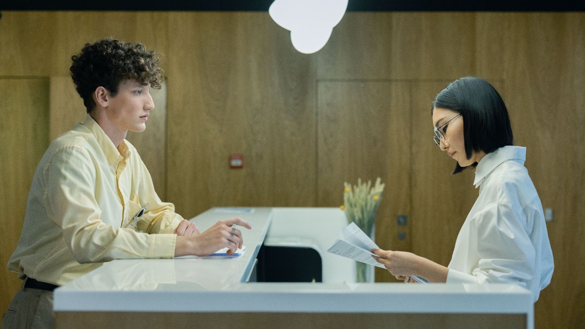 A young man and woman interacting at a modern office reception desk indoors.