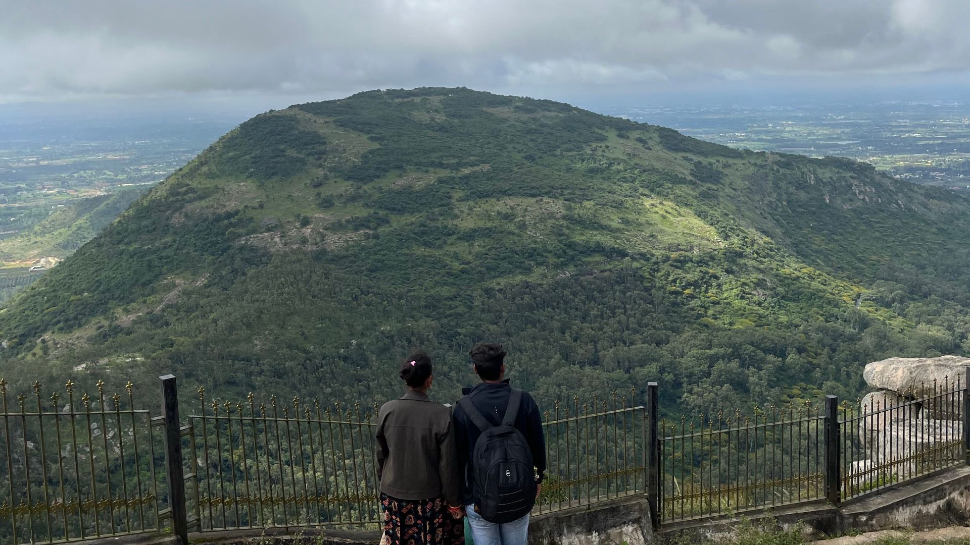 A couple stands on a hilltop overlooking a lush landscape under a cloudy sky.