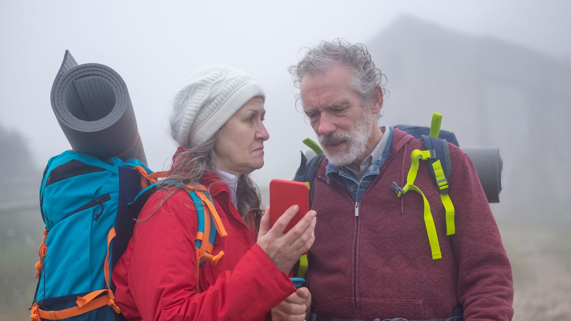 Senior couple with backpacks hiking in a foggy setting, using a smartphone. Travel and adventure lifestyle.