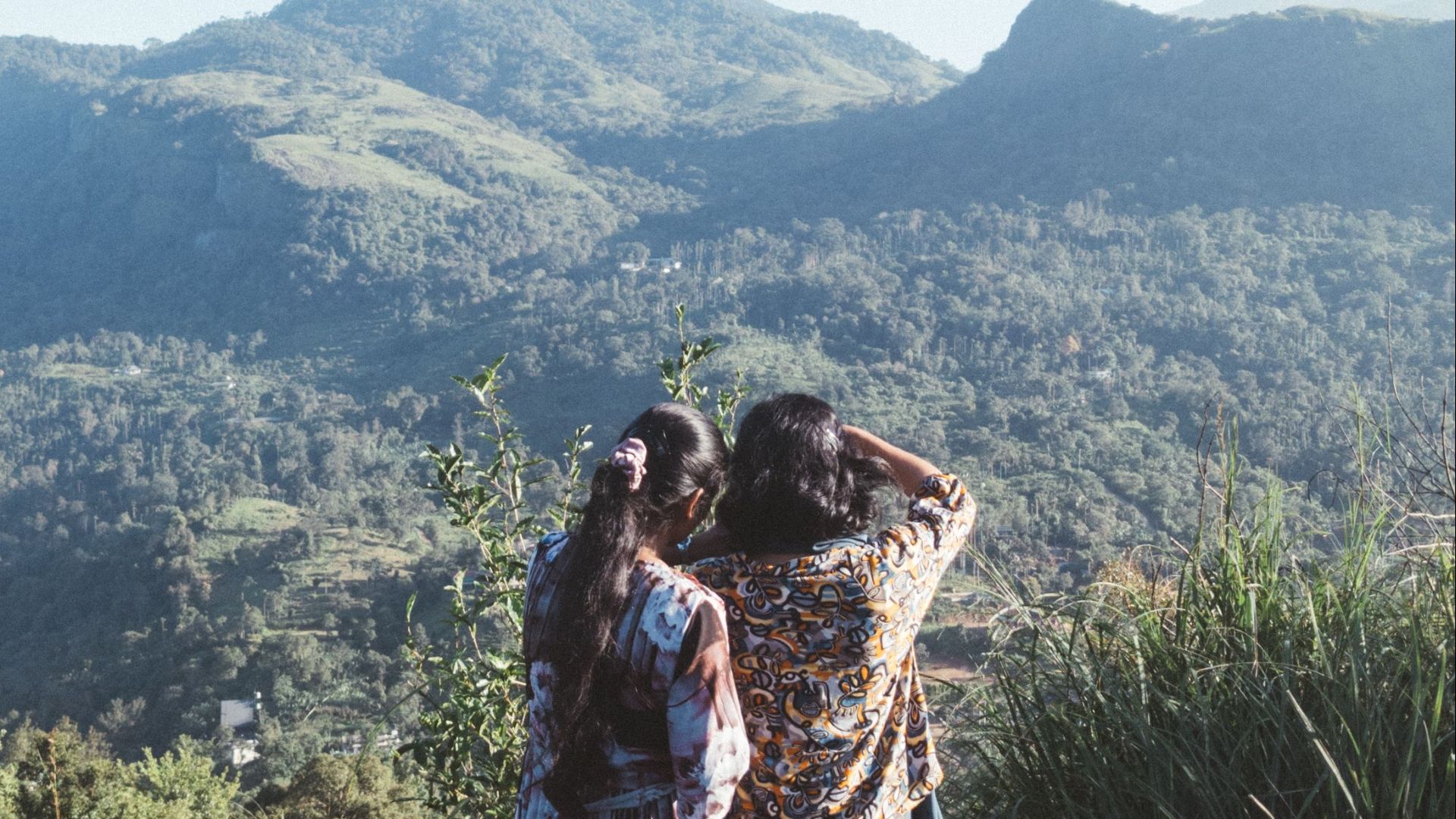 Two women enjoy the scenic view of lush mountains at Kakkadampoyil, India, under a clear blue sky.