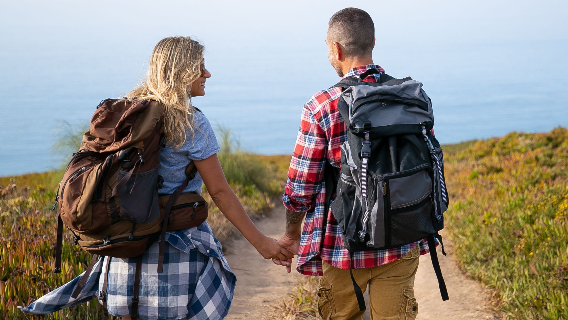 A couple with backpacks walking hand in hand on a trail with a scenic ocean view in Portugal.