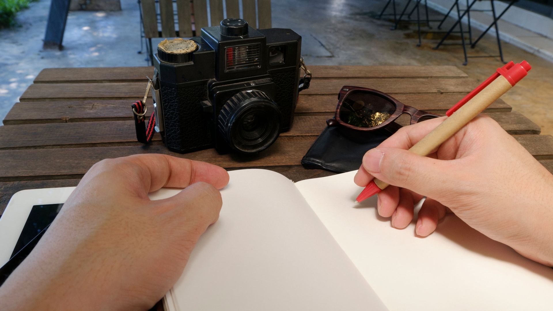 Hands writing in a notebook at an outdoor workspace with a camera and sunglasses.