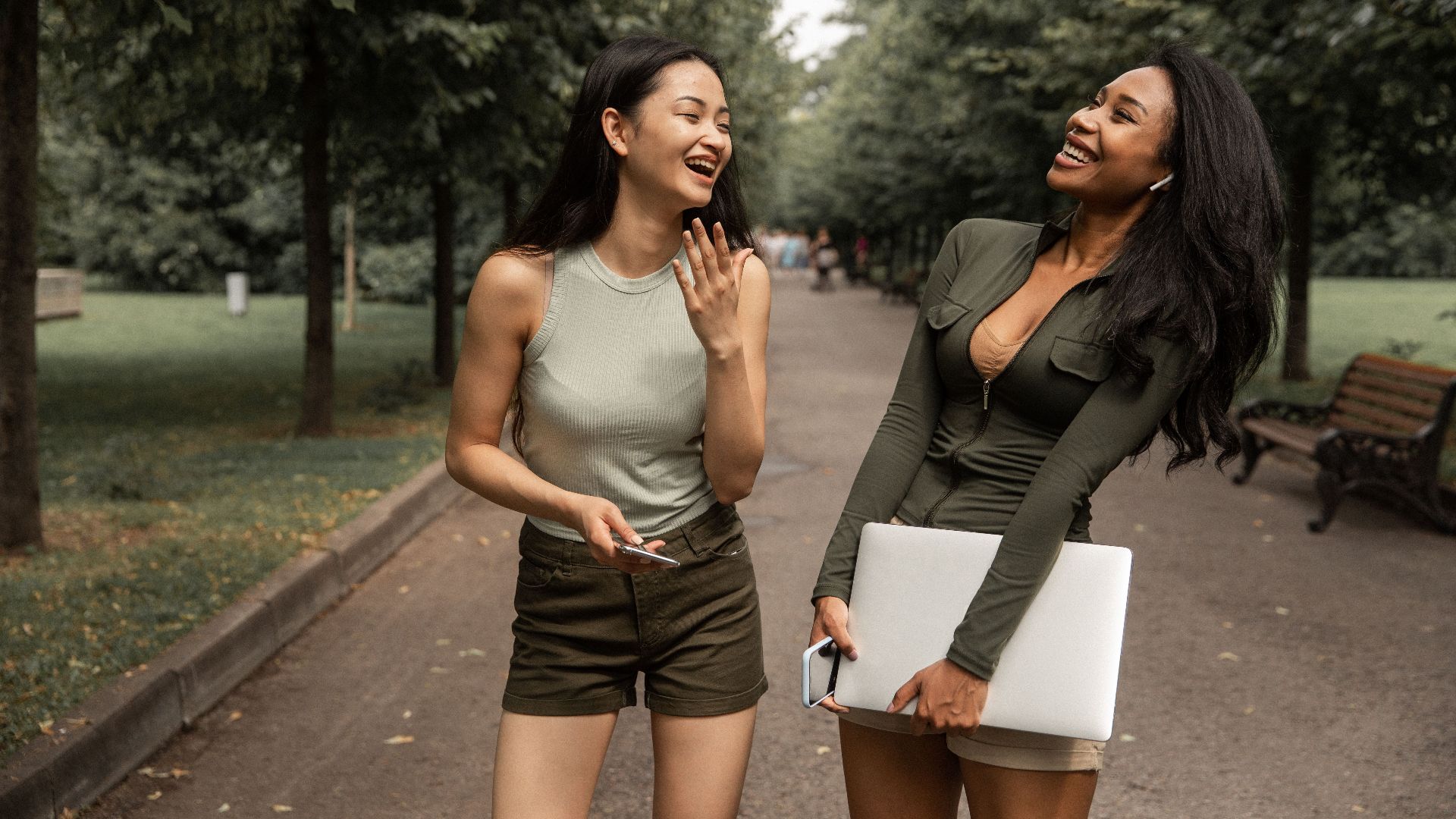 Happy young diverse female friends with smartphone and laptop in hands in casual clothes standing on path amidst lush trees in park and laughing while spending free time together