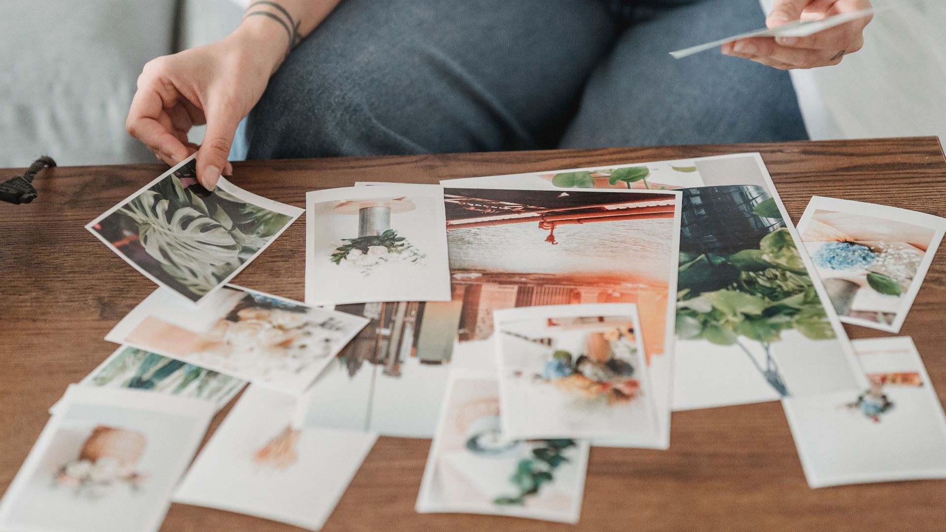 Crop anonymous tattooed female sitting on couch while choosing photos of flowering plants at wooden desk