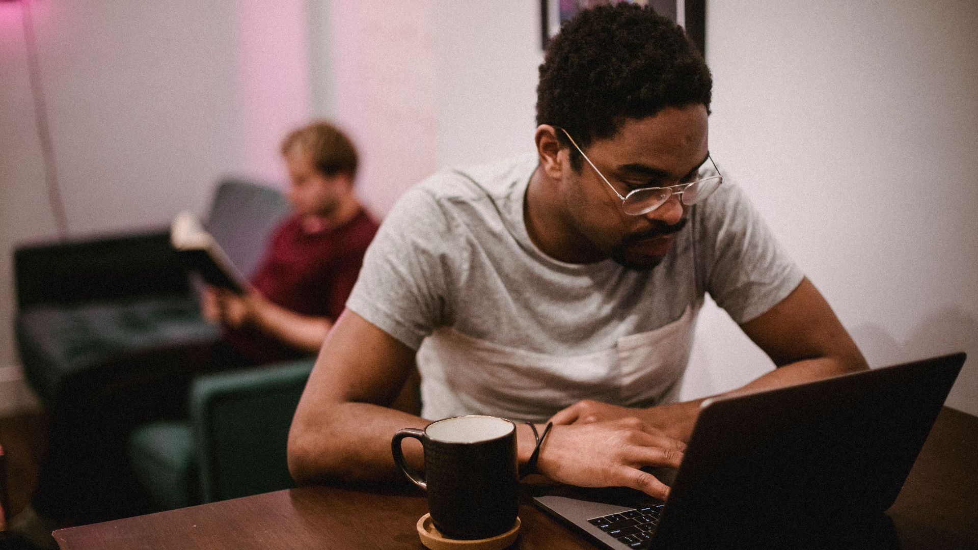 Young man focused on a laptop while another reads in a modern living room.