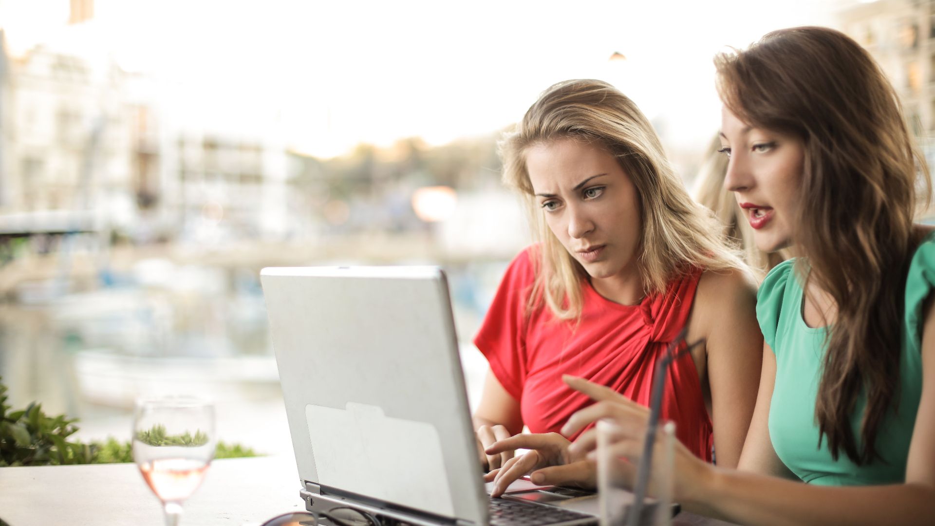 Young female friends in casual wear sitting at table with drinks and using laptop while talking