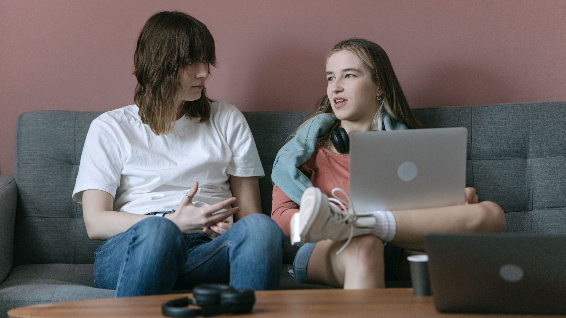 Two young adults engaged in a relaxed conversation on a couch with laptops, depicting a casual indoor setting.