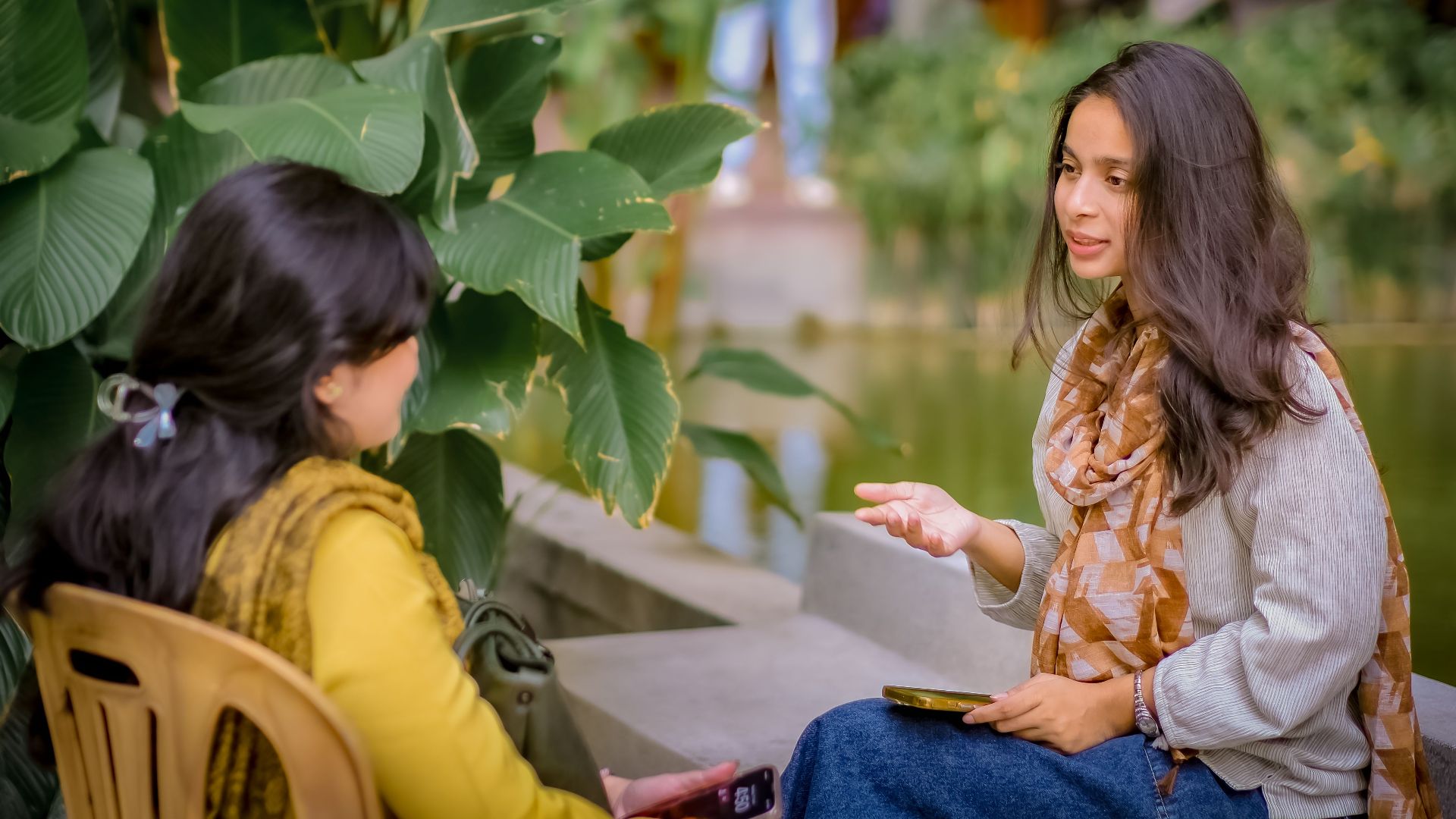 Two women sitting near lush plants having a conversation outdoors. Relaxed and candid atmosphere.