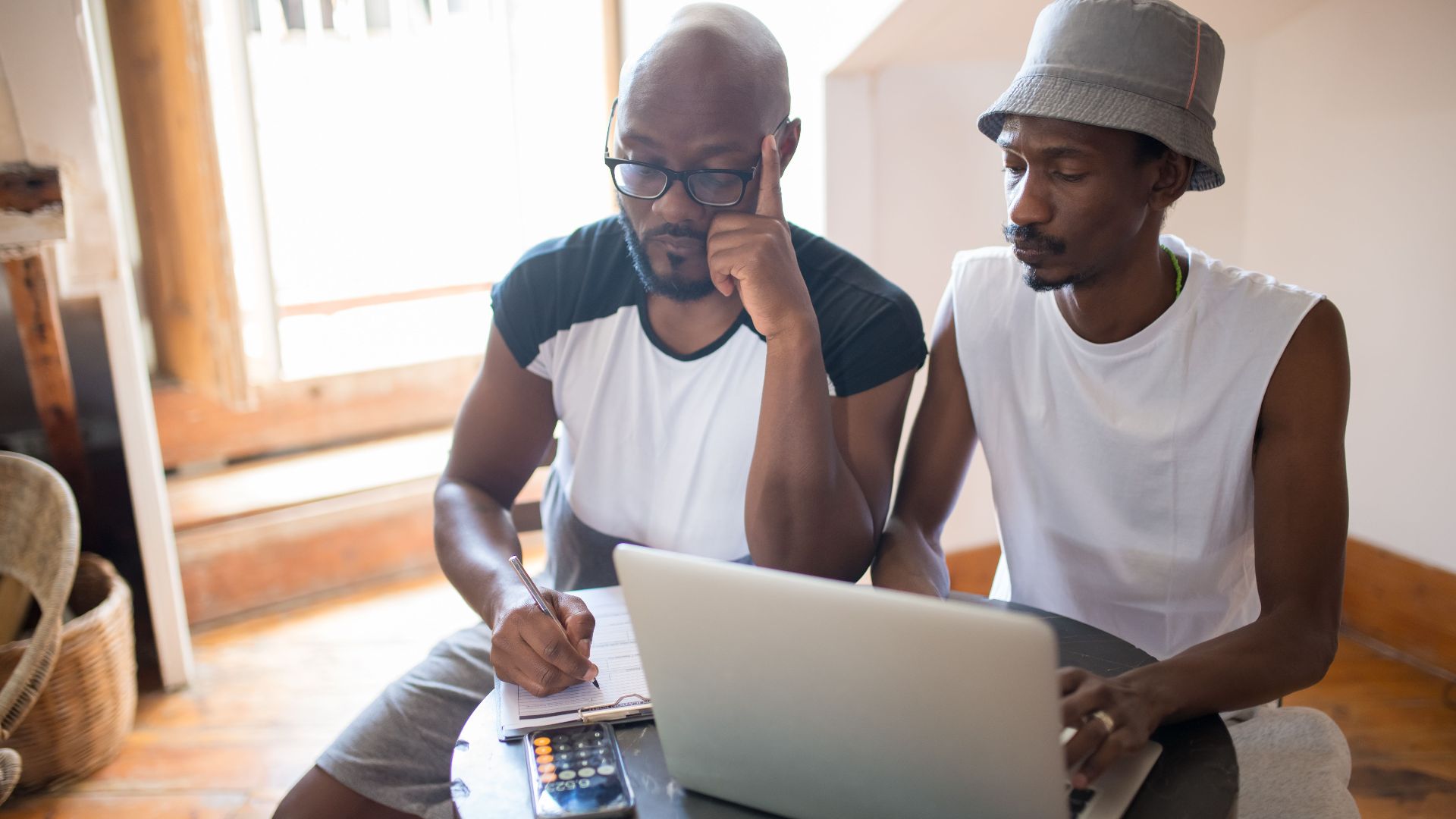 Two men collaborating indoors with a laptop and clipboard, showcasing teamwork and focus.