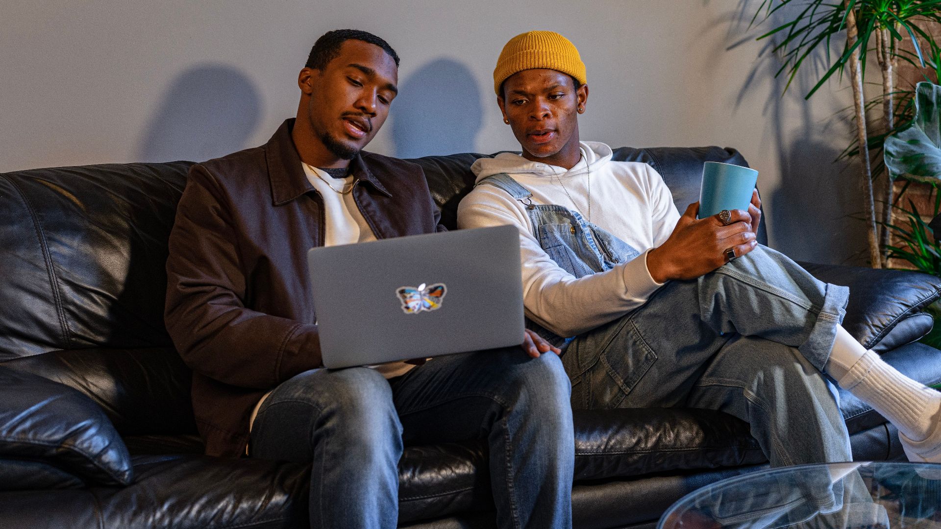 Two men sitting on a sofa using a laptop, enjoying a relaxed indoors setting.