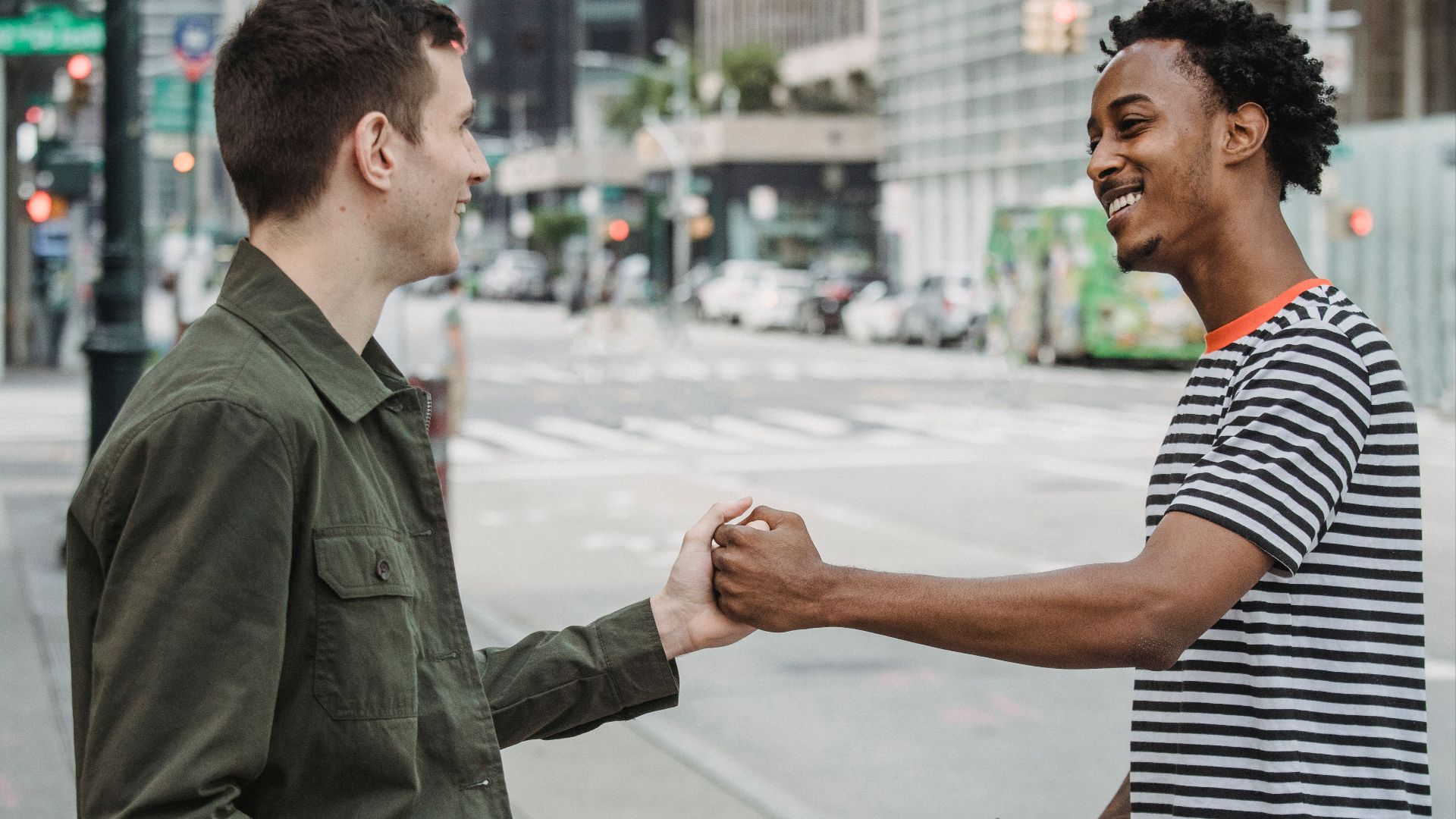 Side view of positive young multiracial male friends in casual clothes smiling and giving dap while greeting each other on city street