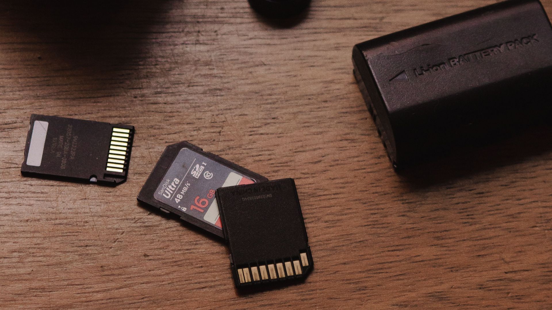 Close-up of camera lenses, SD cards, and battery on wood.