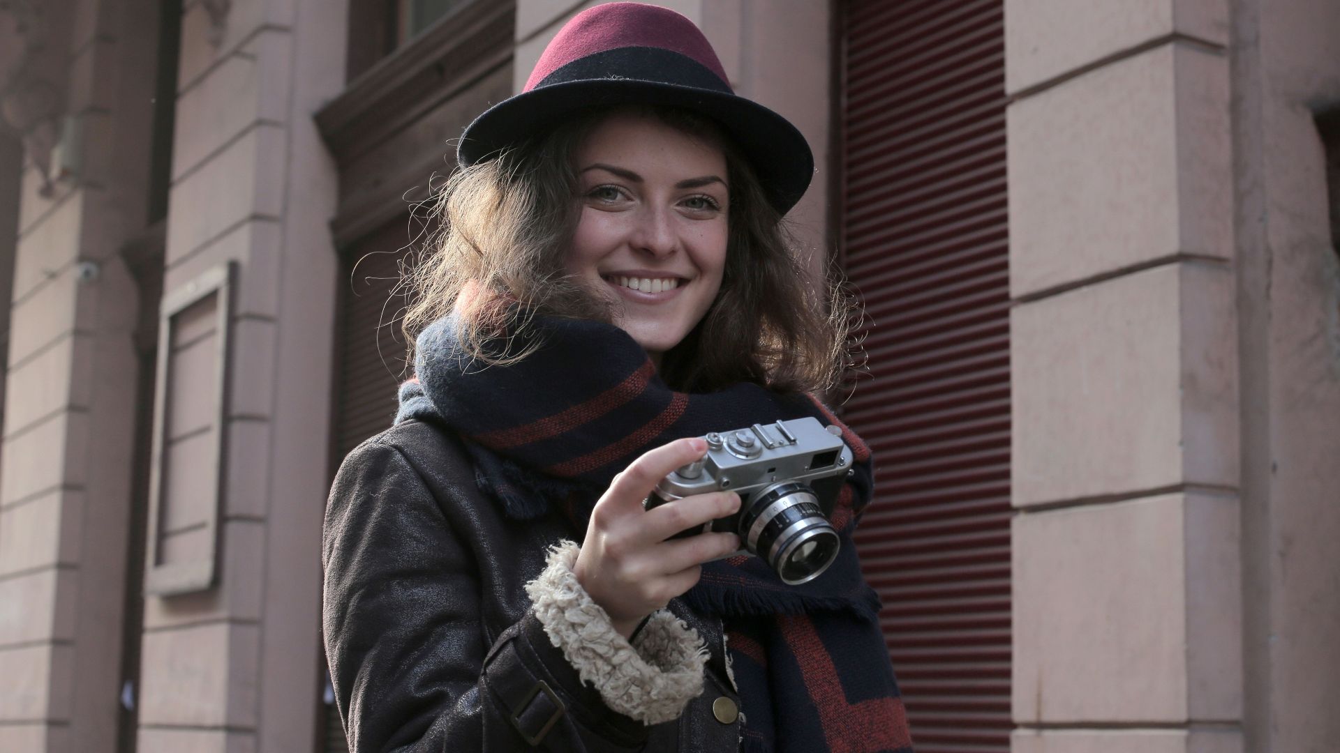 Young woman in winter attire smiling with camera on urban street, exuding style and warmth.
