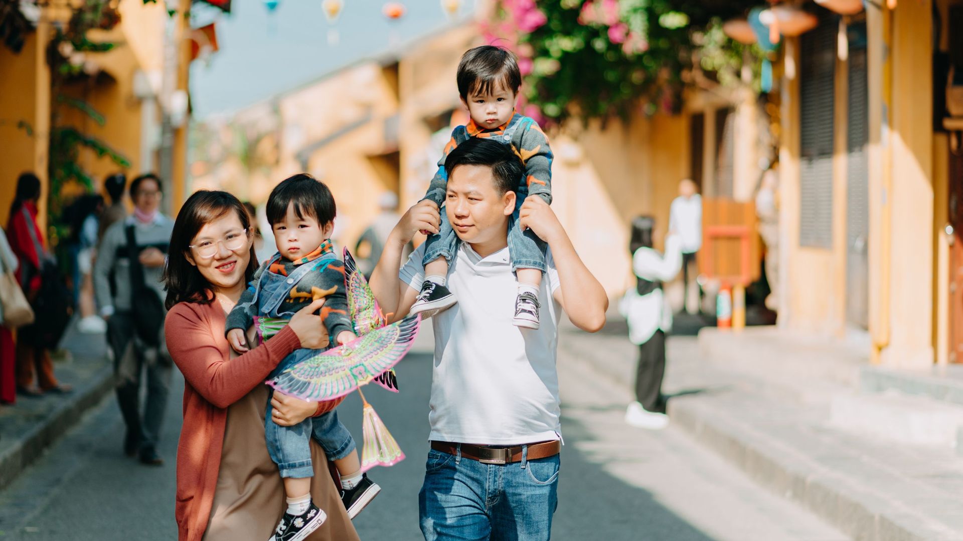 Cheerful Asian parents with sons walking together on asphalt road near buildings with hanging decorations on sunny street of town