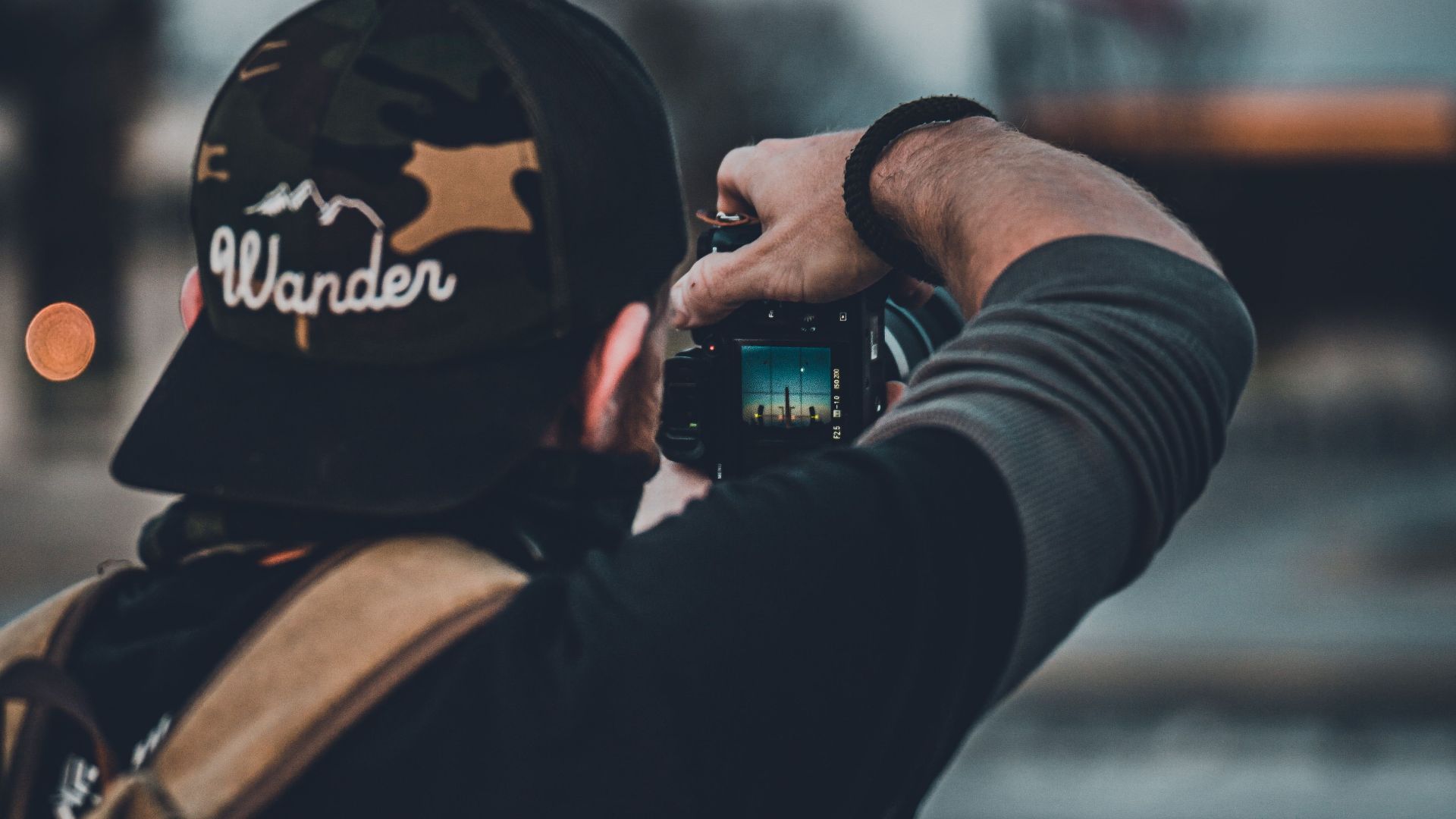 Back view of a photographer with a backpack and cap taking a picture in an outdoor setting.