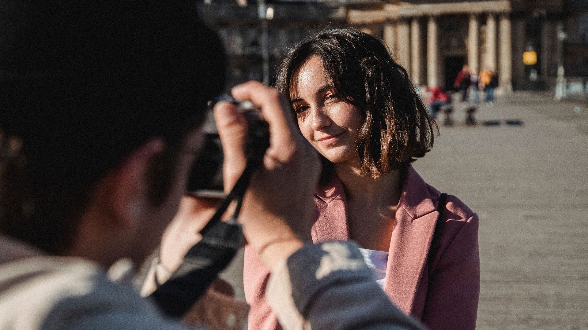 A woman in a pink jacket smiles during an outdoor photoshoot under daylight.