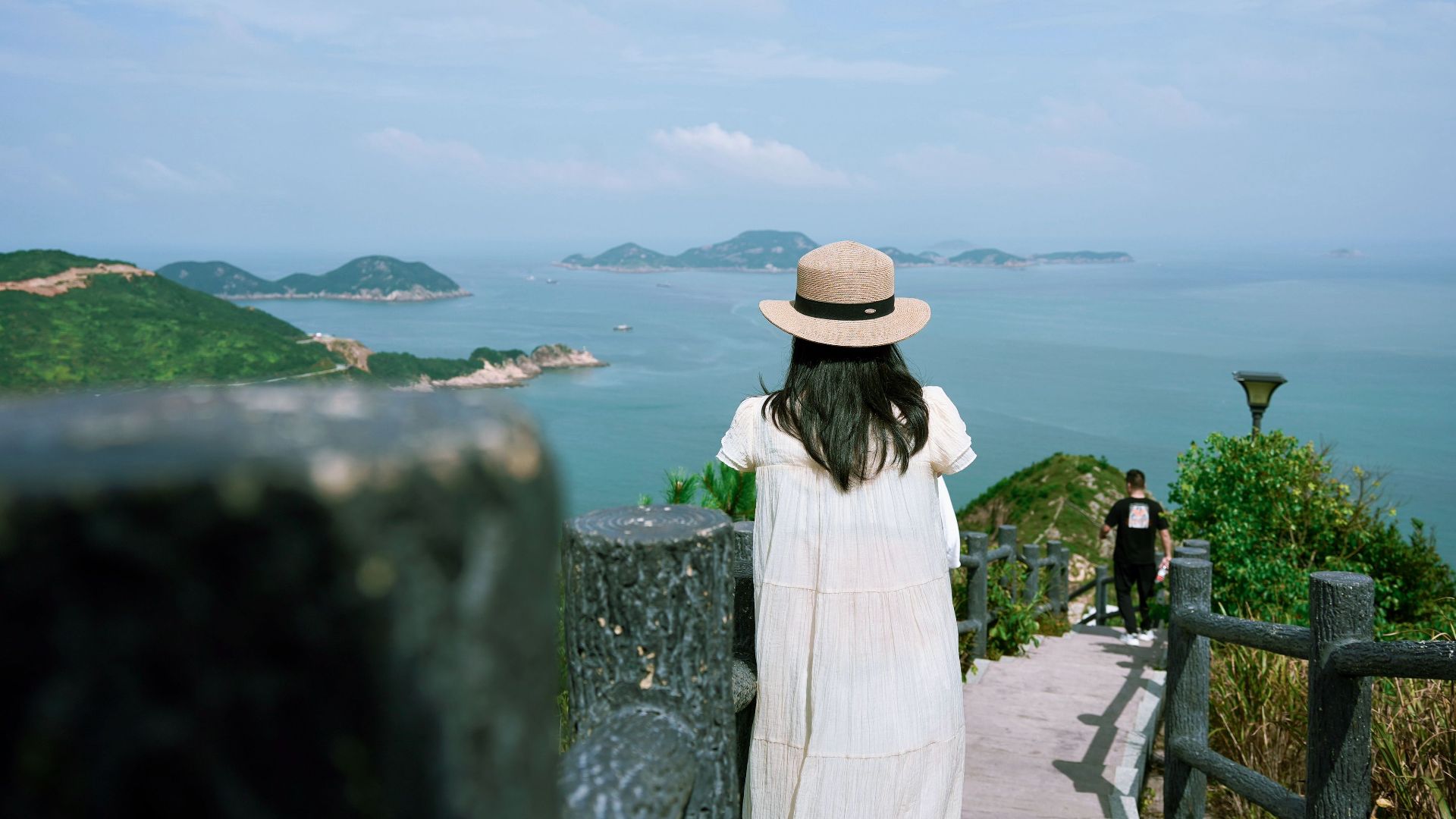 A woman in a hat gazes at the sea and islands from a hilltop path.