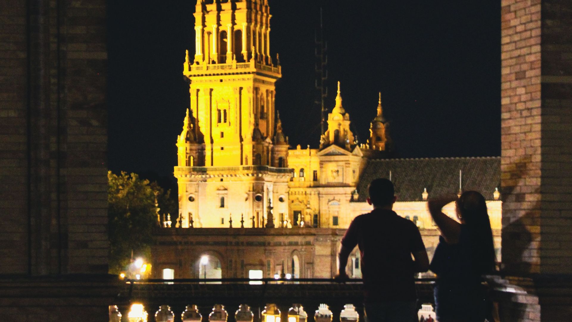 Silhouettes admire the illuminated tower of Plaza de España, captured through an arch in Seville at night.