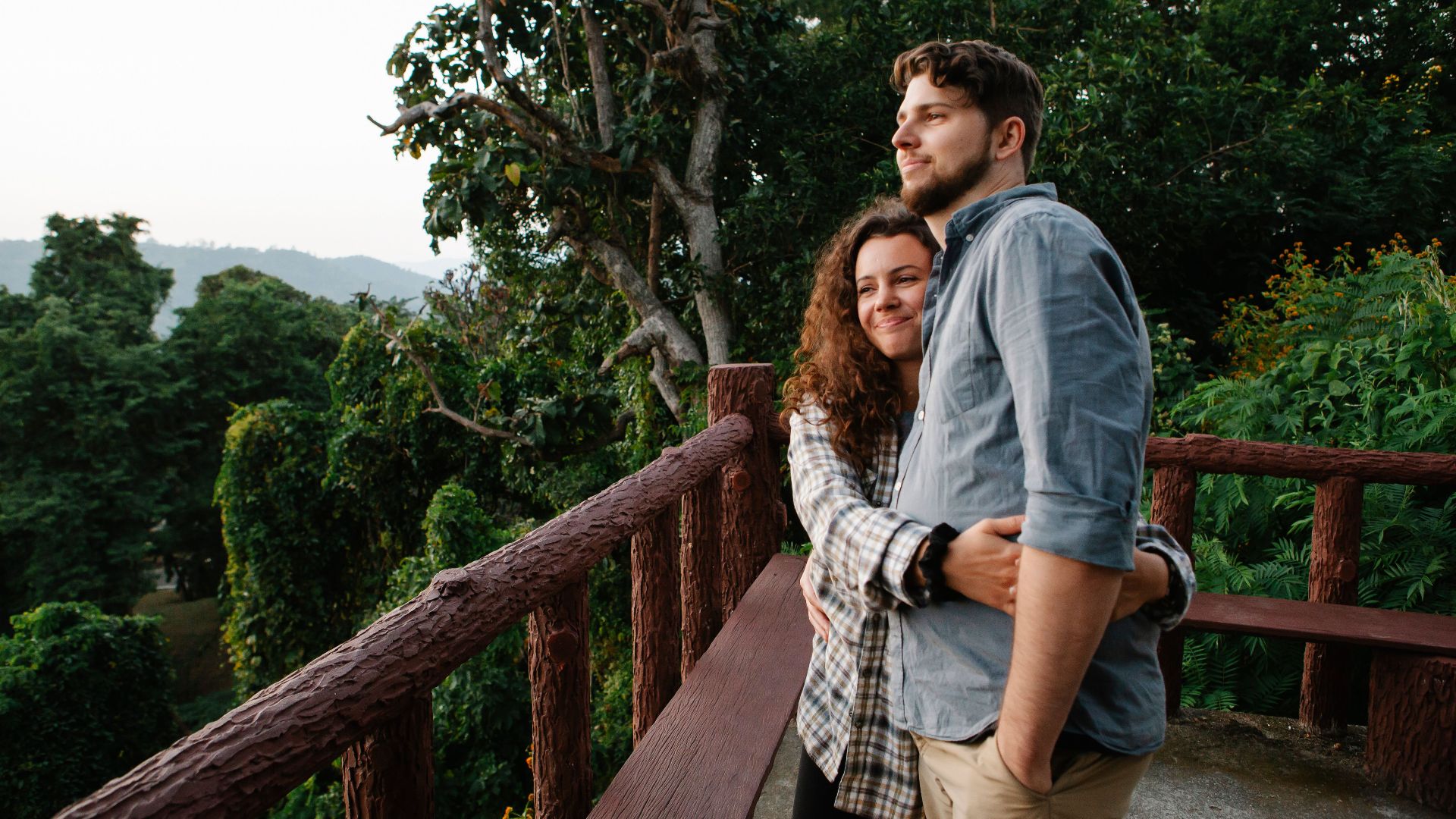 Cheerful young female in casual outfit hugging content boyfriend while admiring together picturesque lush forest standing on wooden observation platform
