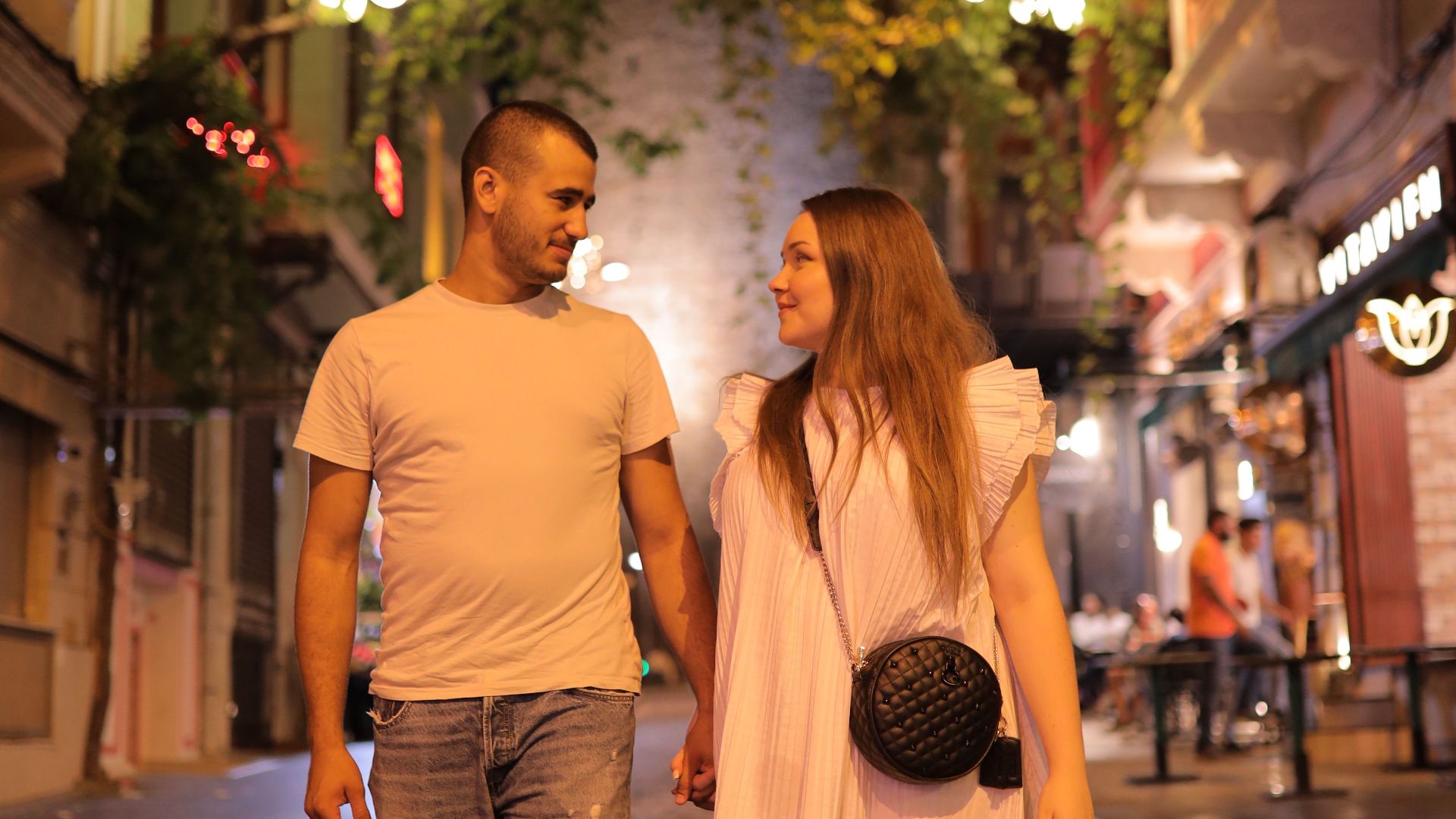 Couple walking hand in hand near illuminated Galata Tower, Istanbul at night.