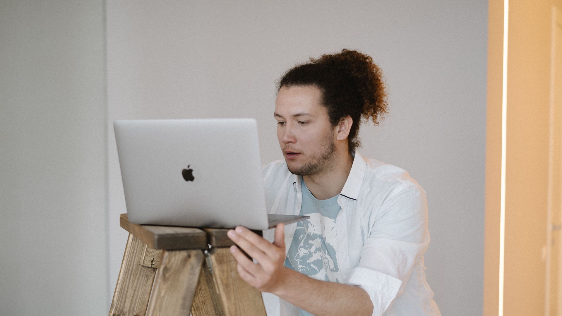 A man using a laptop while standing on a wooden ladder in a minimal indoor setting.