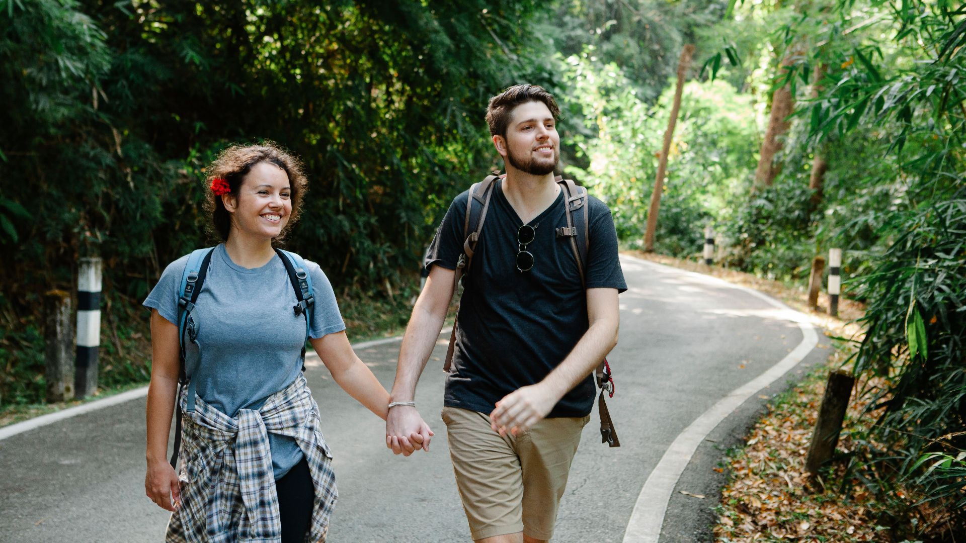 Positive loving young couple in casual outfits and backpacks holding hands while strolling on asphalt road between lush tropical trees during hiking in park