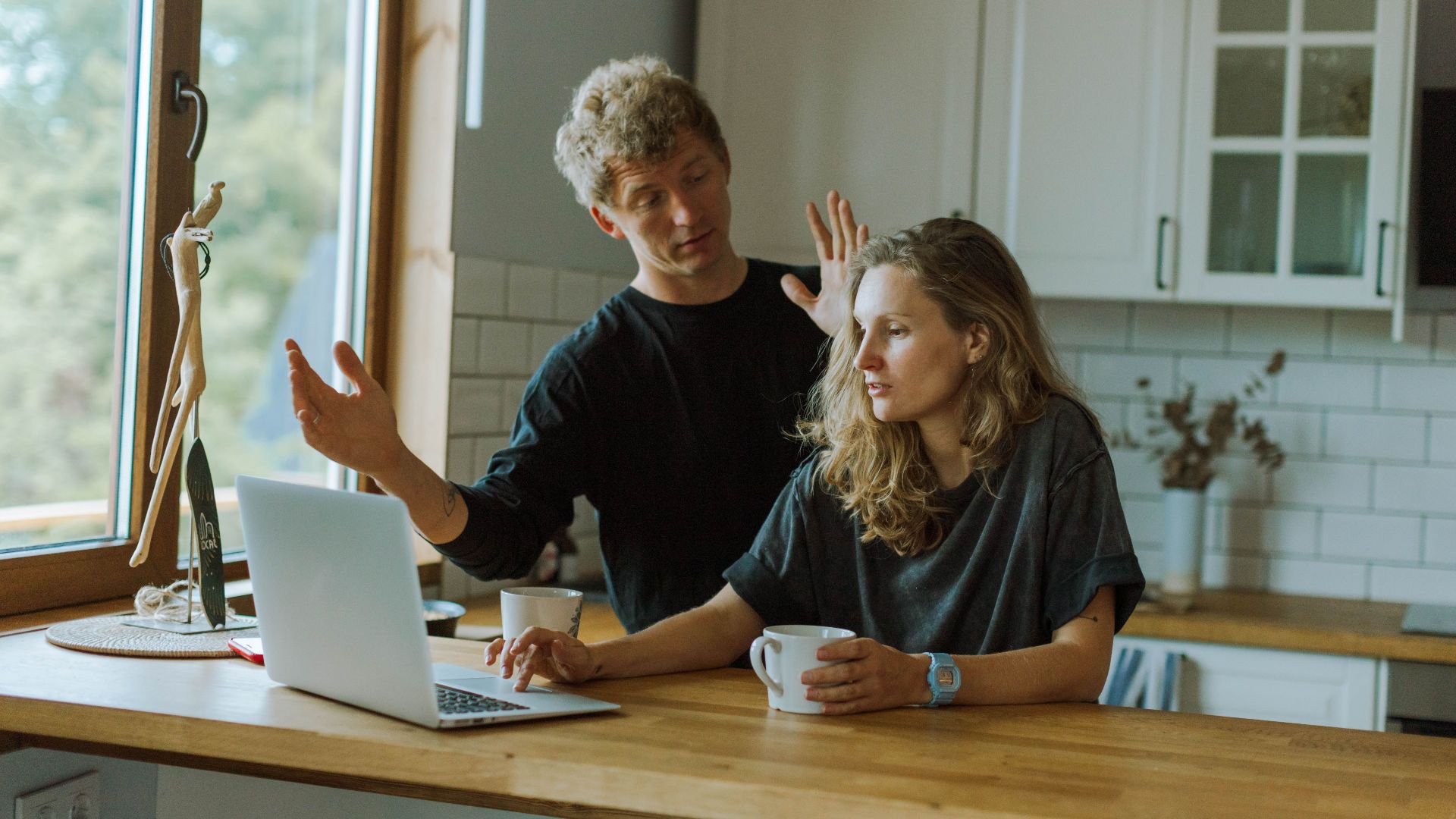 A couple interacting in a bright kitchen using a laptop at a wooden counter.
