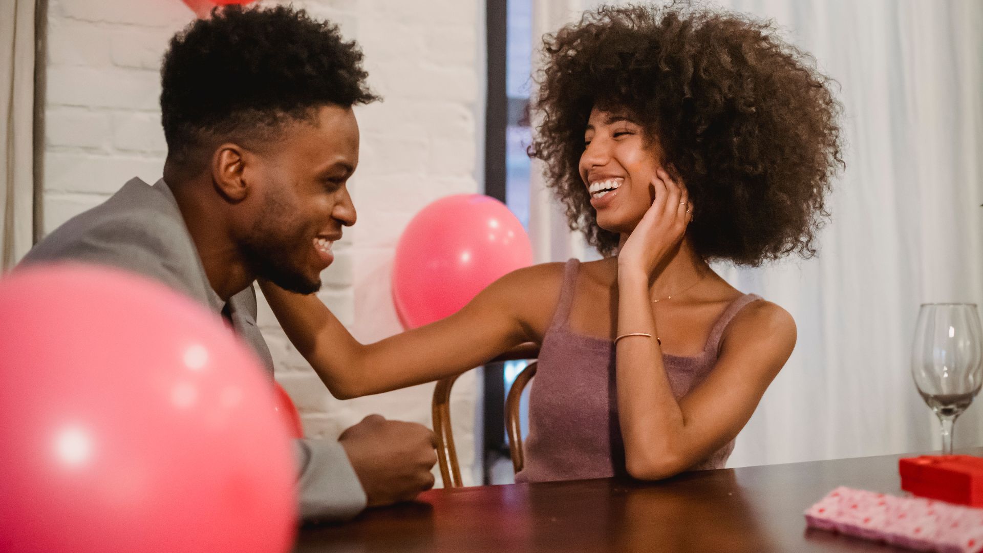 Young black couple in elegant clothes talking and laughing while sitting at table with wineglasses and gifts near balloons at light home with curtains