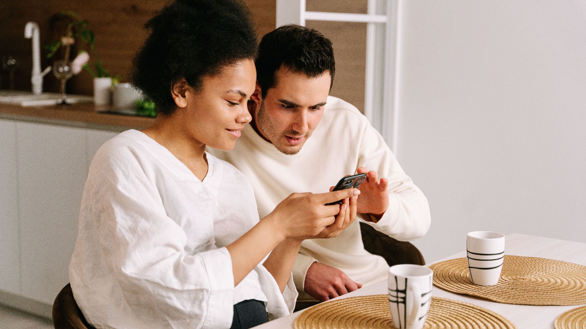 Happy couple sitting in kitchen browsing online shopping on smartphone, sharing moments together.