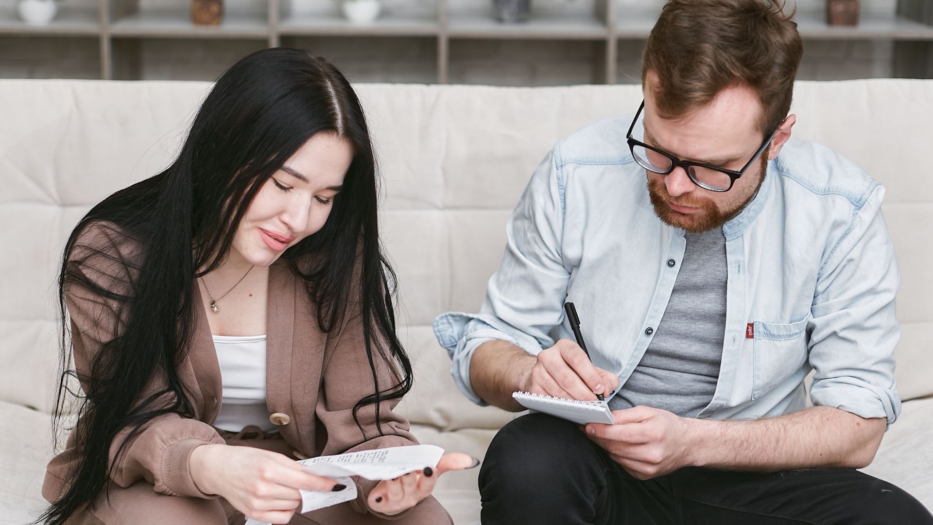 A man and woman sitting indoors, reviewing expenses with notebooks and money on the table.